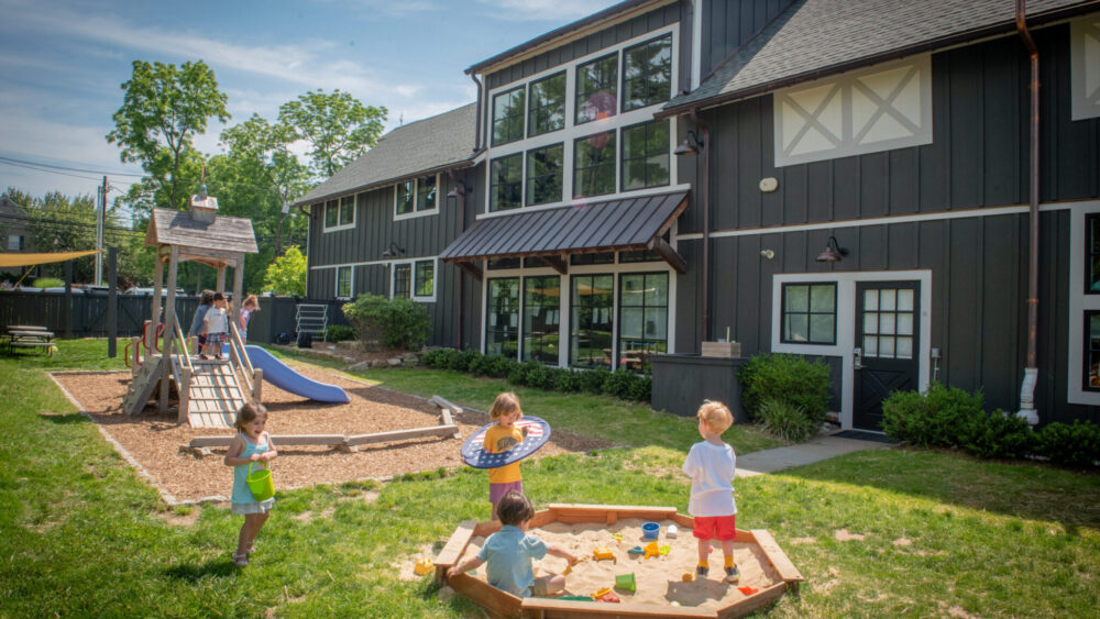Watch "Franchising with Haven" - Our Childcare Franchise Opportunity 7 Children playing outside at Haven Gladstone Playground in New Jersey, featuring a wooden play structure, sandbox, and modern preschool building under a sunny blue sky.